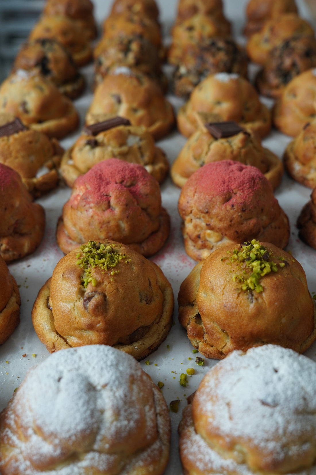 Tray of thick, tall NYC-style bakery cookies with pink, pistachio, powdered sugar, and chocolate chip tops — Baked Up.