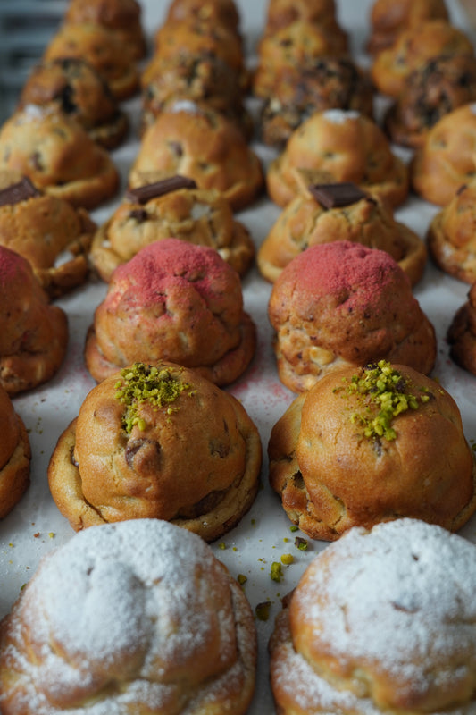 Tray of thick, tall NYC-style bakery cookies with pink, pistachio, powdered sugar, and chocolate chip tops — Baked Up.